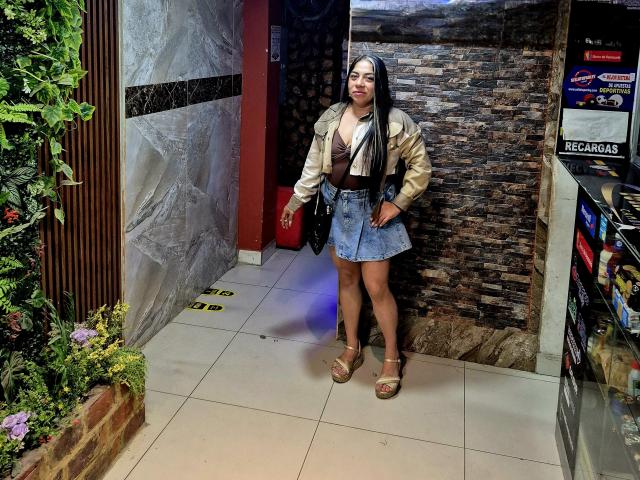 EvaLunaLatina with long black hair, brown skin, wearing a beige jacket, brown bra, denim skirt, brown belt, and tan sandals, standing in a room, holding a black purse.