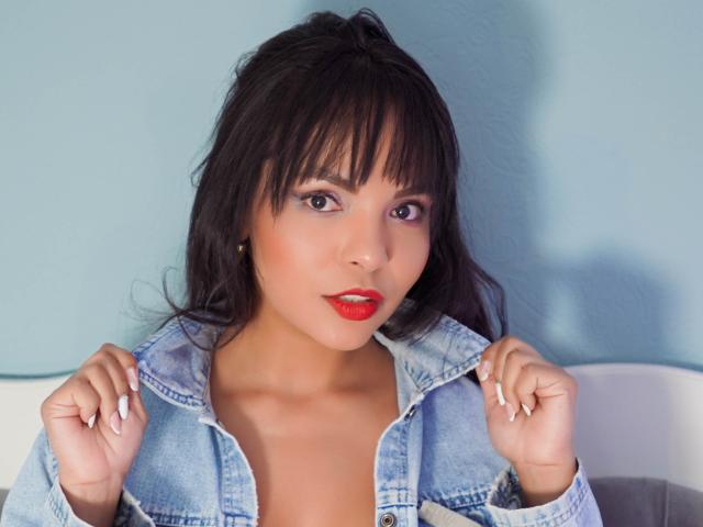 JessicaFiore with dark hair, red lipstick, and blue denim jacket, hands raised, looking at camera, showing her tits slightly. She's got a cute face and a sexy smile.