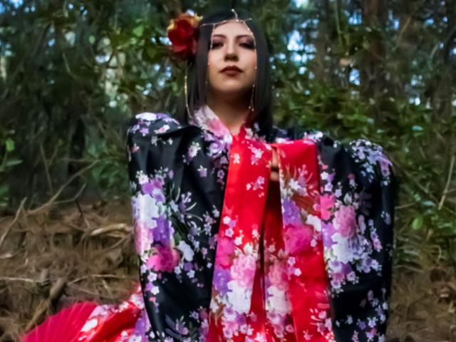 EvangelineAx with dark hair, red flower in hair, wearing black floral kimono with pink and white flowers, red sash, standing in forest, looking confident, holding sash.