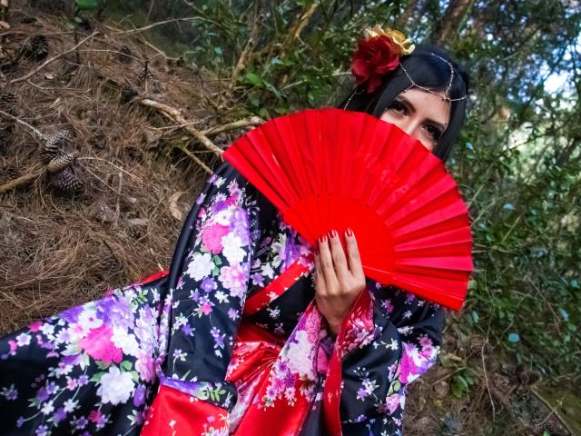 EvangelineAx with floral kimono, red fan covering face, black hair, red flower, and gold hairpiece, holding fan, red nails, standing in forest, showing only eyes and fan.