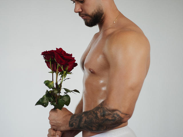 ChrisBlake, sin camisa, sosteniendo una rosa roja, brazo tatuado, piel brillante, collar de oro, pantalones blancos, mirando hacia abajo, perfil lateral, barba corta, fondo neutro..