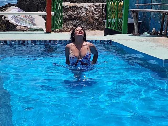 HeraDiosa with medium tits, in a blue pool, wearing a colorful bikini, hands on her ass, smiling, water up to her chest, green door in background.