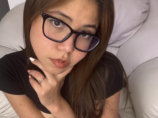 MaddieBlossom has brown eyes, glasses, and pouty lips, wearing a black shirt, with her hand on her neck, showing a ring, and white nails, on a white couch.
