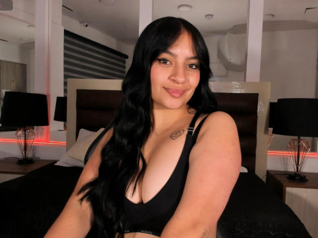 AlexaMiranda with long black hair, smiling, wearing a black bra, showing her tits, small tattoo on right shoulder, in a bedroom with black lamps and red LED lights.