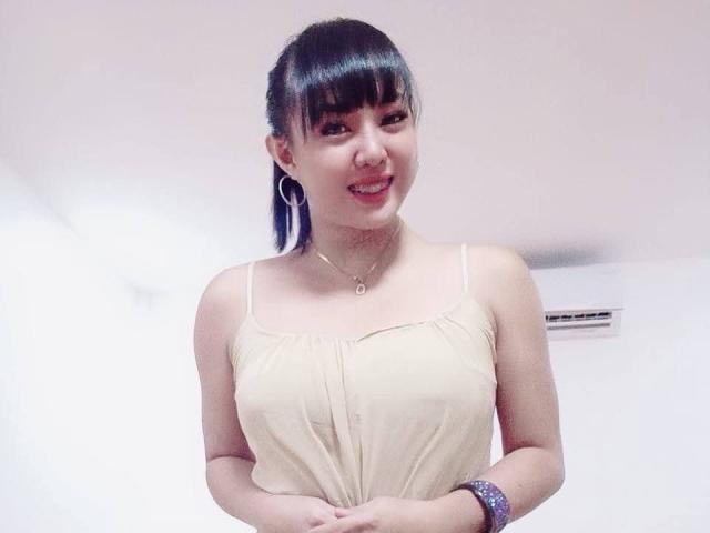 LadyAsh69 with black bangs, blue hair, smiling, wearing a beige spaghetti-strap top, showing small tits, hoop earrings, and a bracelet, in a white room.