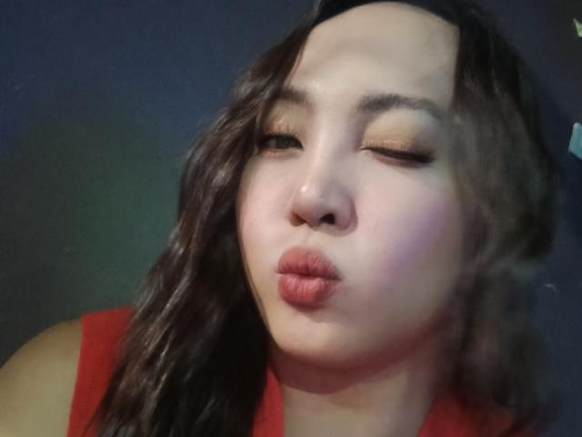 LadyAsh69 with pouty lips, slightly closed eyes, and wavy dark brown hair, wearing a red top, looking relaxed and cute, with a subtle green light in the background.