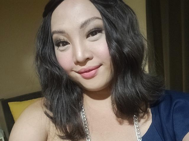 LadyAsh69 with wavy black hair, light skin, and pink lipstick, wearing a navy top with a silver chain, smiling at the camera, showing her tits.