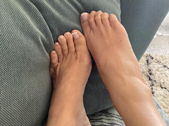 I piedi di mayacorse, con una pelle marrone media, dita leggermente divaricate, sono appoggiati su un divano verde con una texture particolare. L'alluce destro del suo piede si sovrappone al mignolo del piede sinistro..