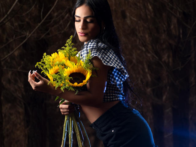 IrinaFerrero with long black hair, checkered shirt, and black pants, holding a sunflower bouquet, slightly leaning forward, eyes closed, smiling softly, dark background, subtle blue light.
