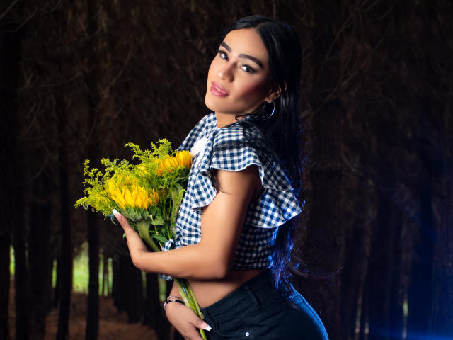 IrinaFerrero with long black hair, wearing a checkered top, holding a bouquet of yellow flowers, looking over her shoulder, smiling, wearing black pants, in a dark forest.