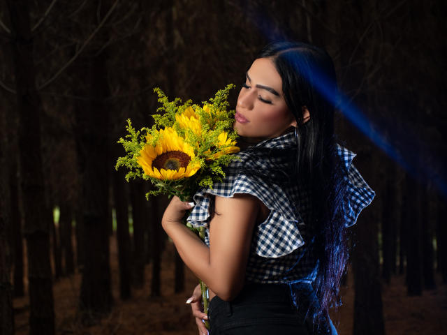 IrinaFerrero with long black hair, checking out a sunflower and yellow greenery, holding them against her chest, wearing a checkered shirt, dark forest background, soft lighting.