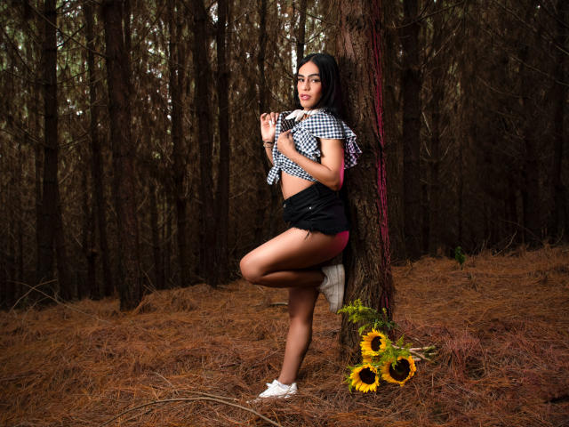 IrinaFerrero with titties in a checkered top, black shorts, and white sneakers, standing against a tree in a forest, leg raised, holding her shirt, with sunflowers at her feet.