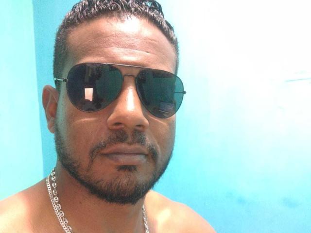 BladeFire with dark sunglasses, short curly hair, and a beard, wearing a silver chain, looking serious, shirtless, against a bright blue background.