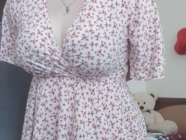 Leiliia's white floral dress with pink flowers, showing her tits, has a deep V-neck and a waist tie. She's wearing a silver necklace. A white teddy is in the background.