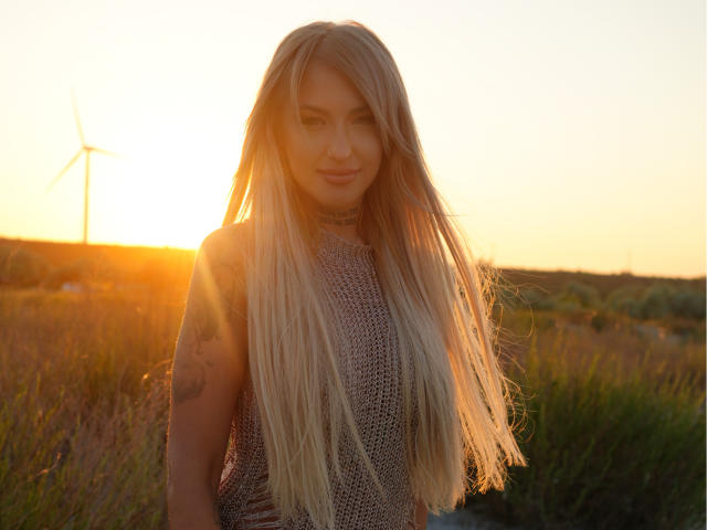SinnersMuse with long blonde hair, wearing a sleeveless knitted top, standing in a field at sunset, sun behind her, wind turbine in the distance, subtle tattoo on her arm.