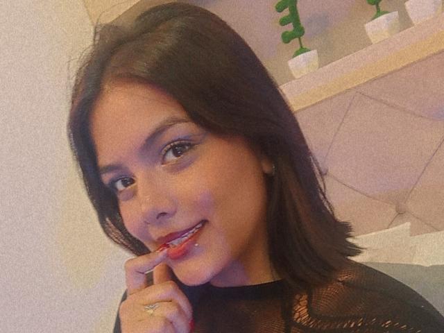 Emily Roberts ha i capelli castano scuro, sta sorridendo con il rossetto rosso, si morde un dito, indossa una maglia nera in rete e ha un anello al dito..