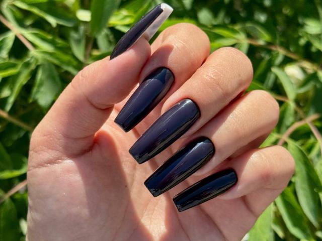 A mão de Skendi, com unhas longas, brilhantes e pretas, exibindo suas unhas impecáveis, sem brilho excessivo, extremamente elegantes e impressionantes, em contraste com um fundo verde e cheio de folhas..