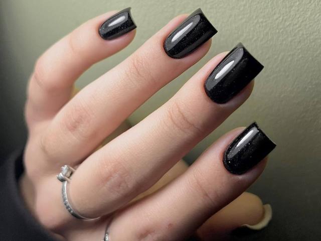 La mano de Skendi, con esmalte de uñas negro brillante y acabado brillante, con un sutil brillo. Lleva un anillo de plata con un pequeño diamante en el dedo índice..
