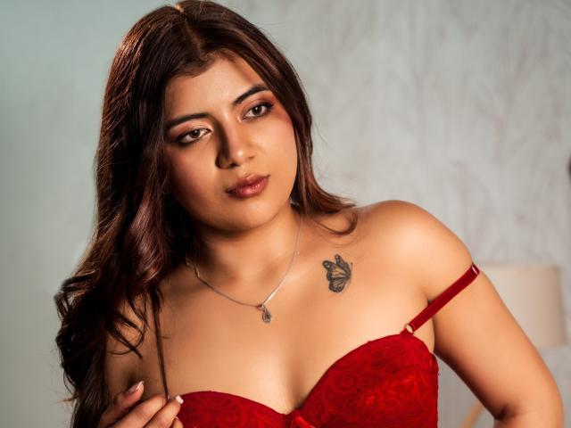 BellaJones69 with brown hair, red lace bra, small tattoo on right shoulder, silver necklace, holding bra strap, looking at camera, slightly parted lips, medium tits.