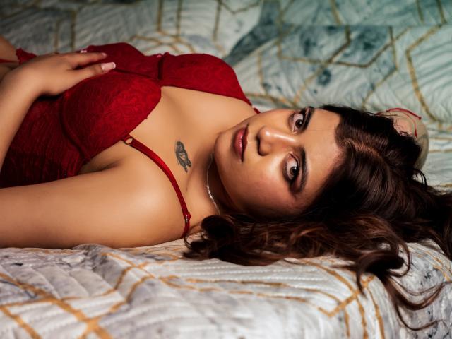 BellaJones69 laying on a bed, wearing a red lace bra, her tits prominently displayed, brown wavy hair, tattoo on her chest, looking seductively at the camera.