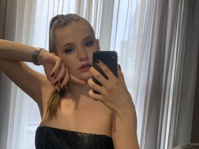IndyVi mit hochgestecktem, braunem Haar, blauen Augen und dunkelroten Nägeln, die ein schwarzes Telefon hält, ein schwarzes Kleid ohne Träger trägt und eine silberne Uhr, und die in die Kamera schaut..