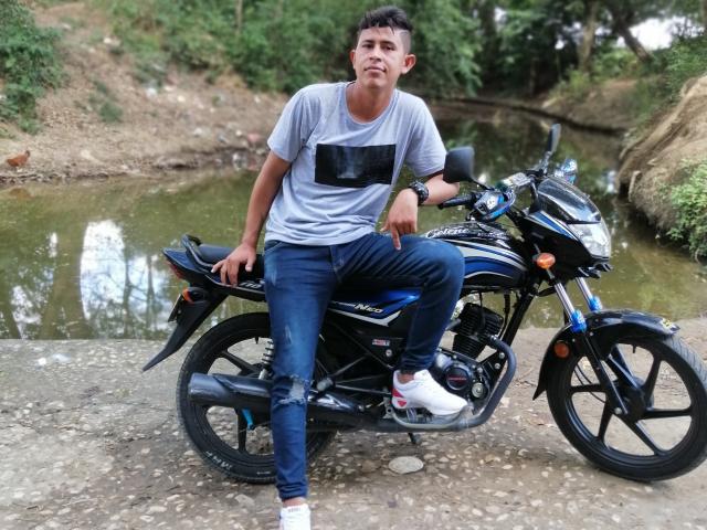 DimyStique, avec du ruban adhésif noir sur la poitrine, assise sur une moto bleue et noire, portant des jeans déchirés et des baskets blanches, près d'un petit étang avec des arbres verts..