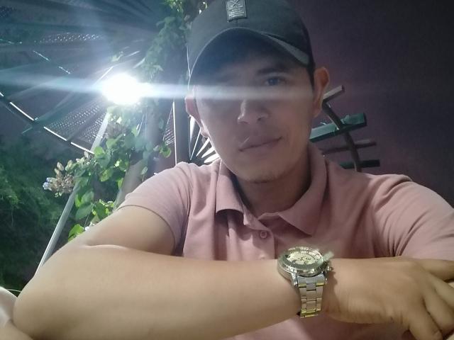 DimyStique, con un tono de piel claro, viste una camisa polo rosa y una gorra negra, tiene un reloj de plata en la muñeca y está sonriendo, con un destello de luz en el fondo..