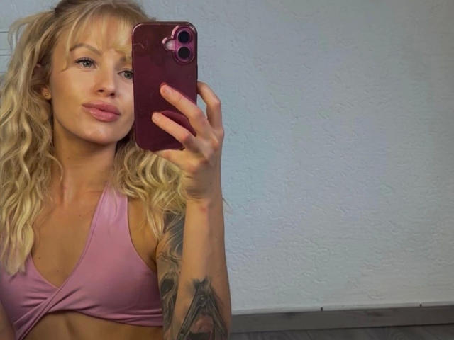 TinyTease-hot with wavy blonde hair, pink sports bra, holding a pink phone, smiling, tattoo on right arm, pink lips, light skin, visible breasts, in a mirror selfie.