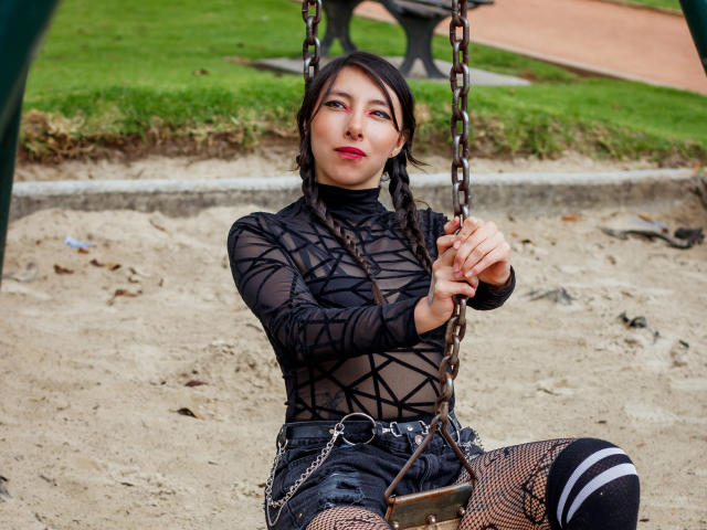 EssledthAnne is sitting on a swing, wearing a black sheer top, fishnet pants, and striped socks. She has braids and is holding the chains with a slightly smug look.