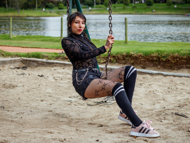 EssledthAnne with taut tits, black mesh top, fishnet pants, black shorts, knee socks, pink sneakers, sitting on a swing, holding chains, in a park, sand ground.