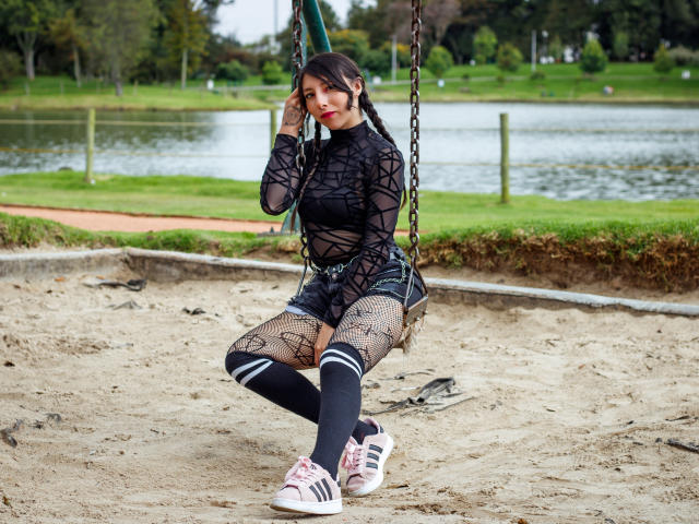 EssledthAnne on a swing, wearing a black, see-through, long-sleeve top, fishnet tights, denim shorts, and pink Adidas sneakers, with her ass slightly raised and her pussy not visible.