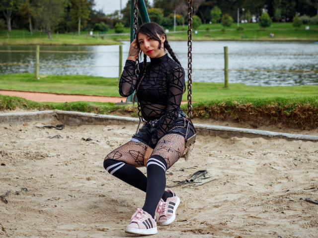 EssledthAnne sits on a swing wearing a black, see-through mesh top, fishnet tights, black shorts, and pink Adidas sneakers. She has black hair and a cute face.