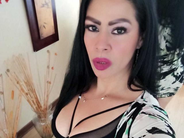 SofiMontes tem cabelo preto, batom rosa e veste uma blusa preta com alças finas que deixam seus seios grandes à mostra. Ela está olhando para a câmera com uma expressão levemente emburrada..