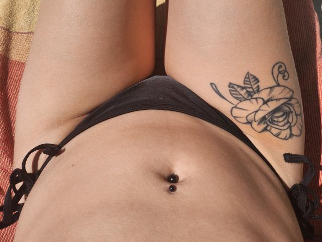 LilouLove's bare midsection, black panties, tattooed thigh with a rose, and a belly button piercing with two black beads. Her skin is light, and she's lying down.