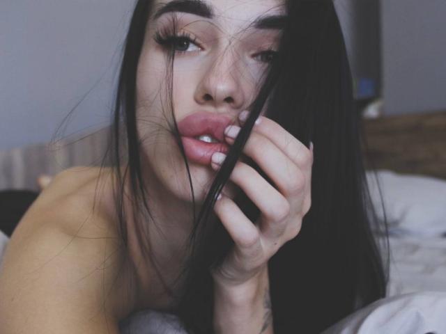 ViktoriaStar with long black hair, biting her finger, showing pink lips, and looking up with half-closed eyes, her hand touching her cheek, on a bed.
