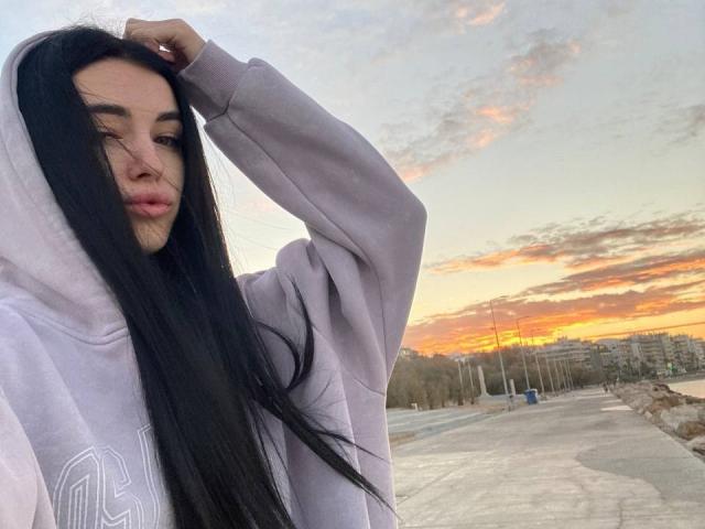 ViktoriaStar with long black hair, pouty lips, wearing a light purple hoodie, hand in hair, sunset sky, orange and pink clouds, empty road, and a few buildings in background.