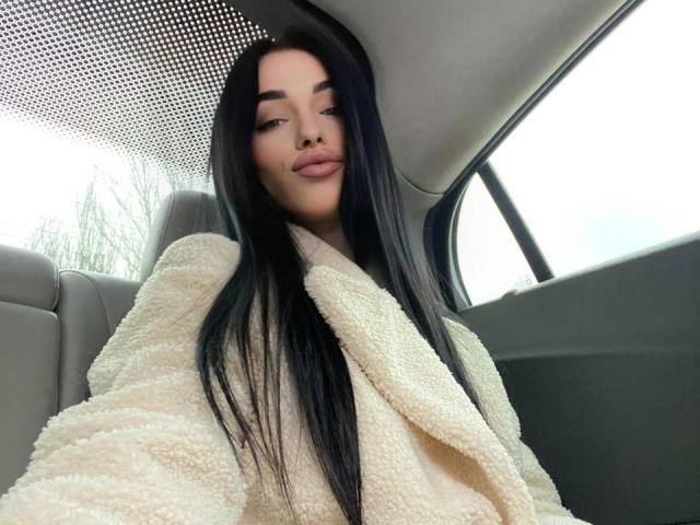 ViktoriaStar with long black hair, wearing a fluffy white coat, sitting in a car, looking down with a slight smile. Boobs not visible, just her face and coat.