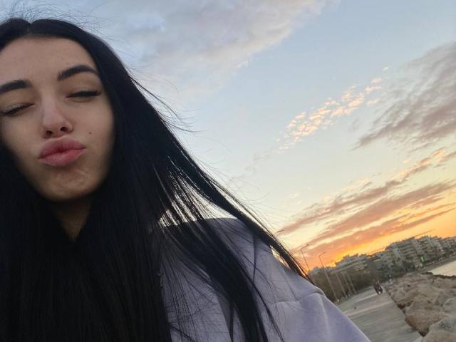 ViktoriaStar has long black hair, closed eyes, and puffy pink lips, wearing a white jacket, with a sunset sky and city in the background.