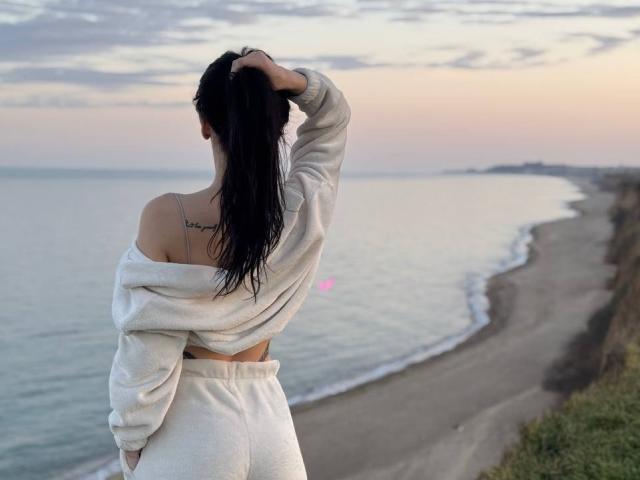 ViktoriaStar with long black hair, off-shoulder white sweater, and matching pants, standing on a beach, hand in hair, showing a small tattoo on her upper back, pussy visible.