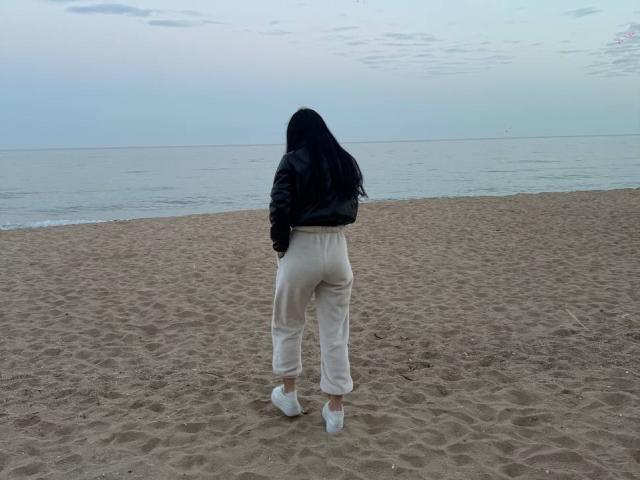 ViktoriaStar with black hair, black jacket, and white pants stands on a sandy beach, facing the ocean, with her ass and white sneakers visible.