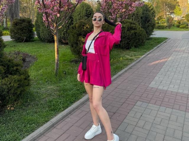 ViktoriaStar with pink dress, white top, white sneakers, black crossbody, holding hair, standing on brick path, green grass, pink magnolia trees, wearing sunglasses, long legs, casual.