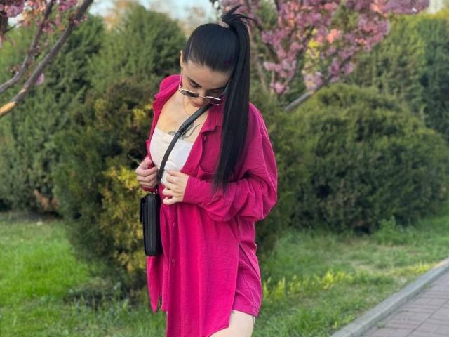 ViktoriaStar with black ponytail, pink dress, white top, sunglasses, black bag, tattoo on chest, standing on grass, holding dress, pink trees in background, green bushes.