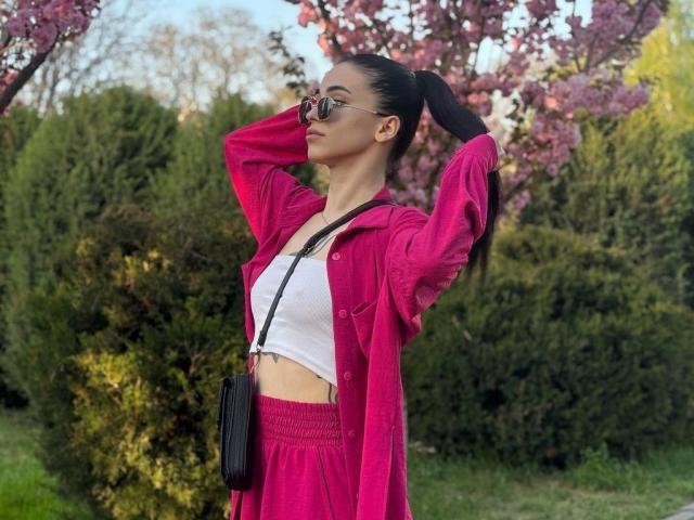 ViktoriaStar with arms up, wearing a pink jacket, white crop top, pink pants, black purse, and sunglasses, standing in a grassy park with cherry blossom trees.