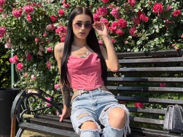 ViktoriaStar with long black hair, pink tube top, ripped blue jeans, tattoos, and sunglasses, sitting on a black bench, hand on face, surrounded by pink roses.