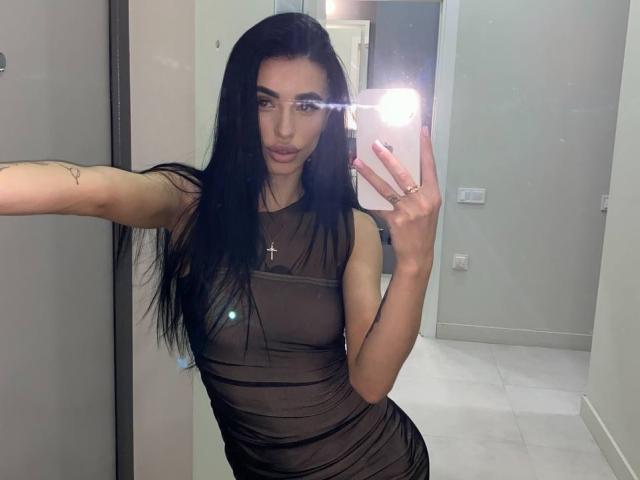 ViktoriaStar with long black hair, wearing a sheer black dress, holding a phone with a flash, showing her small tits, cross necklace, and a tattoo on her arm.
