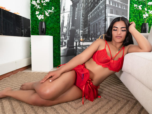 Emmaly Morgan, con i capelli scuri, un reggiseno di pizzo rosso e una gonna rossa, sdraiata sul tappeto, con una mano sulla testa, che appare rilassata, gambe scoperte, seno di media grandezza, carina, attraente, sexy, pelle marrone..
