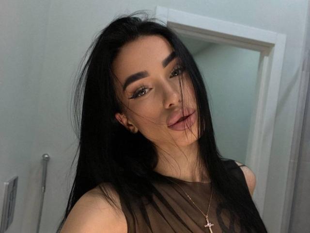 ViktoriaStar has long black hair, dark eyebrows, and full pink lips. She's wearing a sheer black top with a cross necklace, looking at the camera with a slight smile.