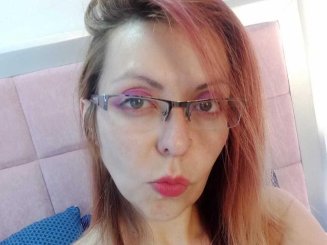 ArtemisFlyve with pink eyeshadow, red lipstick, and glasses, pouting her pink lips, has light brown hair with pink highlights, and is wearing a pink headboard in the background.