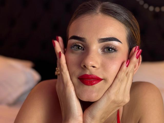 IsabellaMichelle heeft rode lippenstift, donkere wenkbrauwen en rode nagels, met haar handen voor haar gezicht, waarbij ze haar borsten laat zien en een ring draagt aan haar rechterhand. Haar groene ogen zijn gecentreerd..