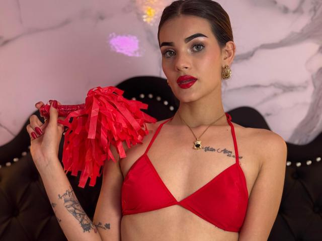 IsabellaMichelle mit rotem BH, die rote Pompons hält, hat Tattoos auf Brust und Arm, trägt goldene Ohrringe und roten Lippenstift, braune Haare im Pferdeschwanz..
